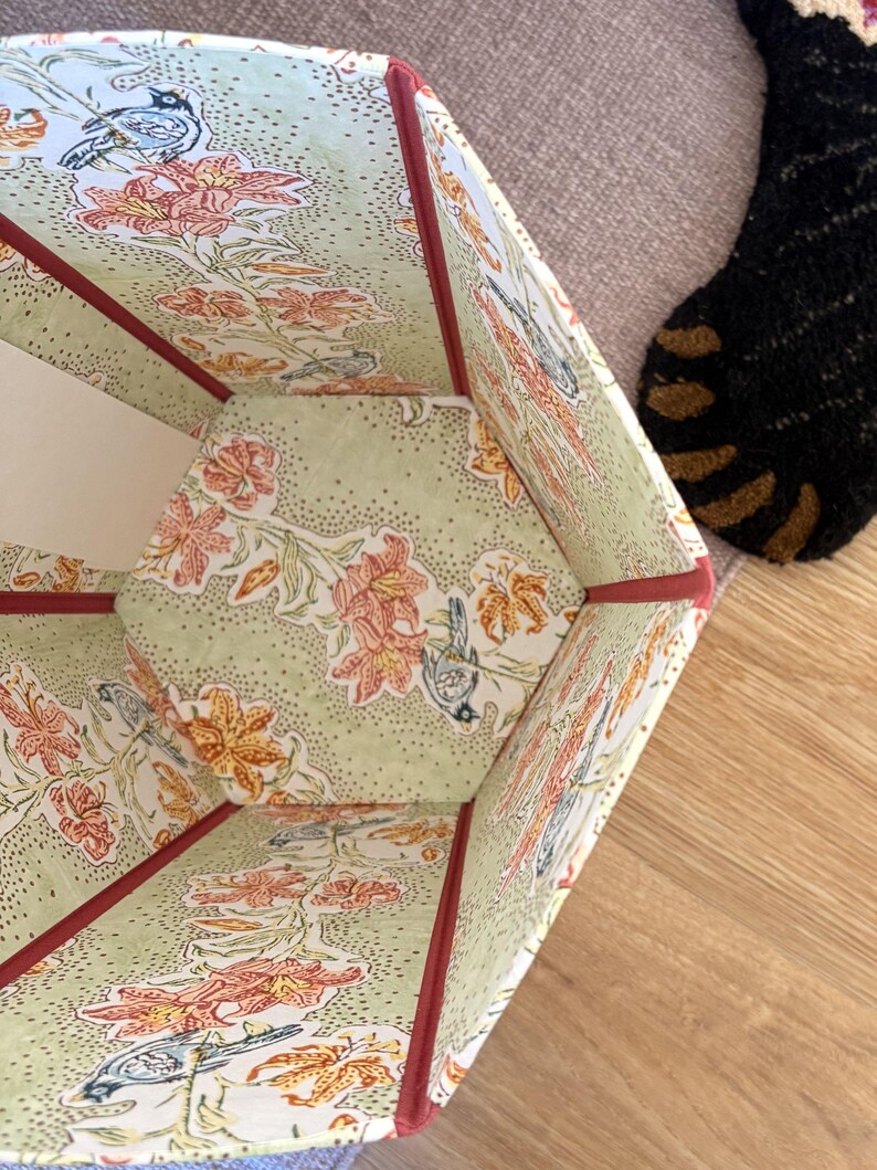 May include: A hexagonal storage box with a floral and bird pattern in shades of orange, green, and white. The edges are trimmed with a dark red binding. The interior and exterior feature the same design.