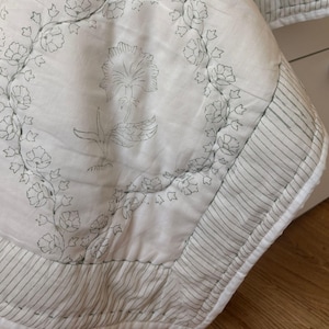 Op de afbeelding: Een witte doorgestikte deken met een grijs bloemenpatroon, voorzien van een centraal bloemmotief omringd door een cirkelvormige rank van kleinere bloesems. De rand bestaat uit grijs en wit gestreepte stof met zichtbare stiksels over het geheel.