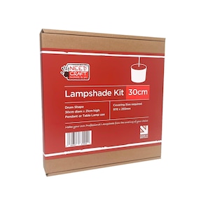 Op de afbeelding: Een vierkante kartonnen doos voor een Needcraft lampenkapset. Het rode label bevat witte tekst met de tekst Lampshade Kit 30cm, Drum Shape, 30cm diam x 21cm high, Pendant or Table Lamp use, en Covering Size required: 970 x 255mm, met een streepjescode.