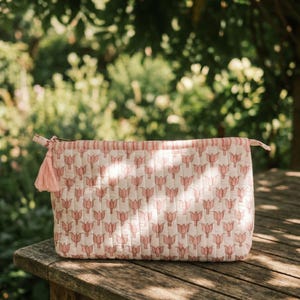 May include: A rectangular, quilted cosmetic bag with a repeating pink tulip pattern on a cream background. The top edge features pink and white stripes, a pink tassel, and a zipper closure. The bag is resting on a wooden surface.