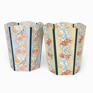 Pode incluir: Duas lixeiras decorativas com estampas florais. Uma é azul clara com flores rosa e laranja, pássaros e listras azul marinho. A outra é amarela com flores laranja, pássaros e listras azul marinho. Ambas têm bordas onduladas.