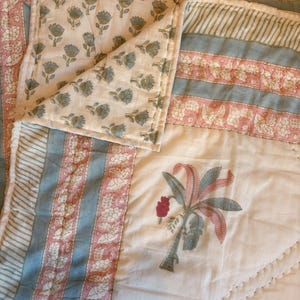 Op de afbeelding: Een gewatteerde textiel met een crèmekleurige achtergrond met een herhalend bloemmotief in blauwgroen. De rand heeft strepen met blauwe en roze bloemmotieven. Een palmboomontwerp is op de quilt gedrukt.