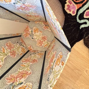 May include: An open, multi-sided lampshade with a floral pattern in orange, pink, and green on a light blue background. The shade has a dark blue trim and is set on a wooden floor. A patterned rug is visible in the background.