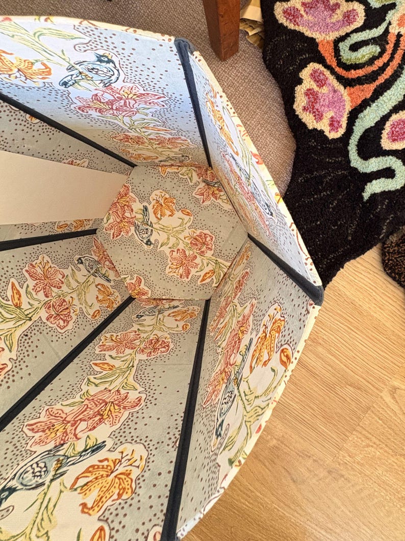 May include: An open, multi-sided lampshade with a floral pattern in orange, pink, and green on a light blue background. The shade has a dark blue trim and is set on a wooden floor. A patterned rug is visible in the background.
