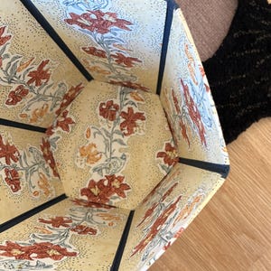 May include: Hexagonal storage container with a floral pattern. The interior and exterior feature a cream-colored background with a repeating pattern of red and orange lilies and green leaves. The edges are trimmed with navy blue fabric.