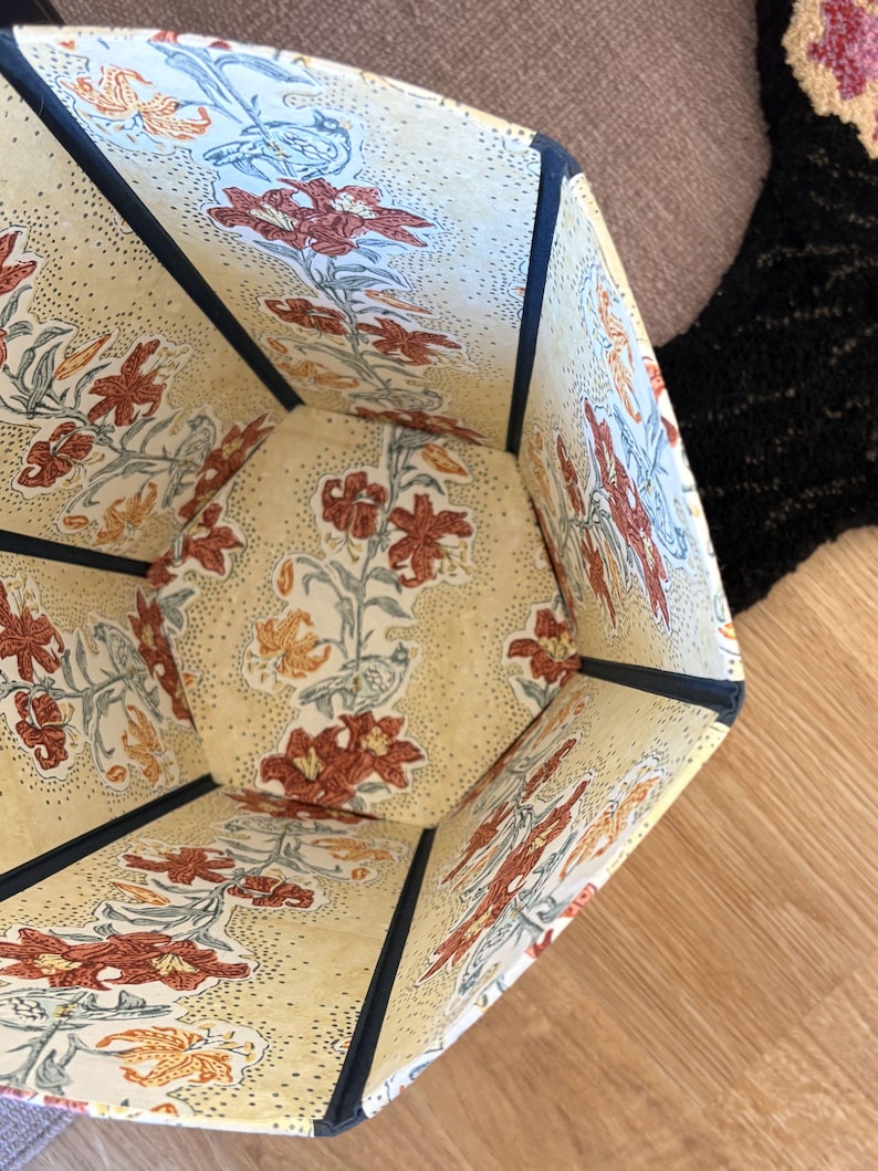 May include: Hexagonal storage container with a floral pattern. The interior and exterior feature a cream-colored background with a repeating pattern of red and orange lilies and green leaves. The edges are trimmed with navy blue fabric.