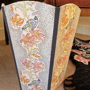 May include: A decorative wastebasket with a floral and bird pattern. The design features orange and pink lilies, green stems, and small blue birds against a light blue and dotted background. The edges are trimmed with a dark gray material.