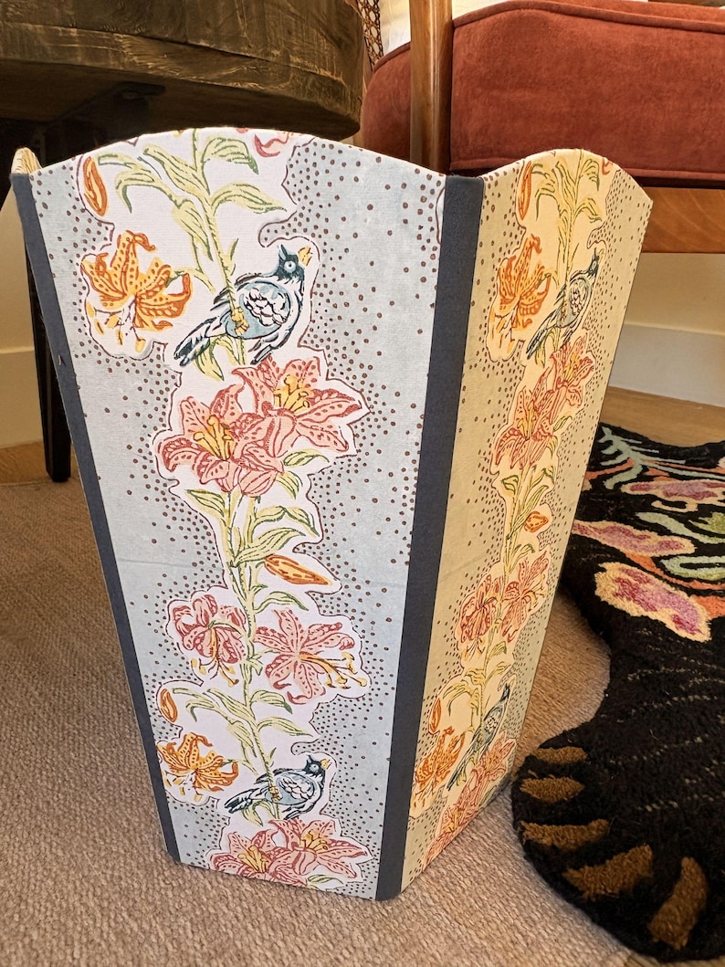 May include: A decorative wastebasket with a floral and bird pattern. The design features orange and pink lilies, green stems, and small blue birds against a light blue and dotted background. The edges are trimmed with a dark gray material.