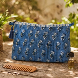 May include: A blue quilted cosmetic bag with a floral pattern, a wooden comb, and a small glass bottle with a dark cap are on a wooden surface. The bag has a tassel and a striped zipper detail.