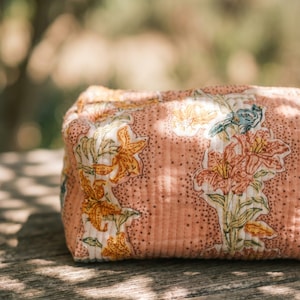 May include: A rectangular, quilted cosmetic bag with a floral pattern. The bag features a peach-colored background with a repeating pattern of orange, yellow, and blue flowers. The bag is sitting on a wooden surface.