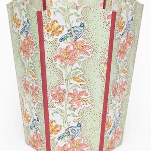 May include: A decorative wastebasket with a floral and bird pattern. The wastebasket features a light green background with a repeating pattern of orange and yellow lilies, blue birds, and red vertical stripes.