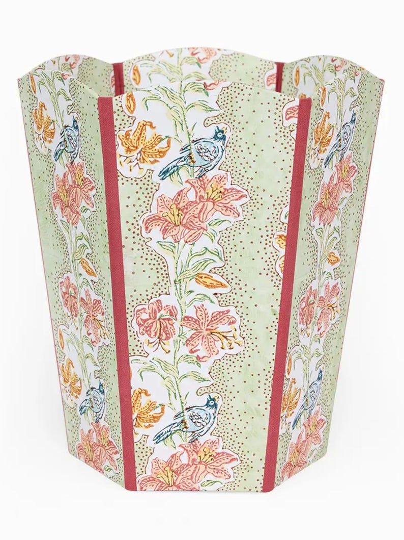 May include: A decorative wastebasket with a floral and bird pattern. The wastebasket features a light green background with a repeating pattern of orange and yellow lilies, blue birds, and red vertical stripes.