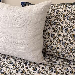 Puede incluir: Primer plano de dos cojines decorativos. Uno es blanco con un diseño floral en relieve. El otro cojín tiene un fondo blanco con un estampado floral azul y amarillo. Los cojines están sobre una cama con sábanas estampadas a juego.