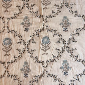 Könnte beinhalten: Ein cremefarbener Stoff mit einem sich wiederholenden Muster aus blauen und grauen Blumenmotiven, die in einem Gitter aus dunkelgrauen Ranken angeordnet sind. Der Stoff enthält abwechselnd große und kleine Blumen und eine gestreifte Kante.