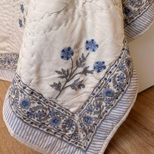 Peut inclure: Un textile matelassé blanc orné d'un motif floral bleu avec des détails cousus à la main. La bordure présente un motif rayé bleu et blanc répété ainsi qu'un motif floral, créant un bord décoratif sur la surface en tissu blanc cassé.