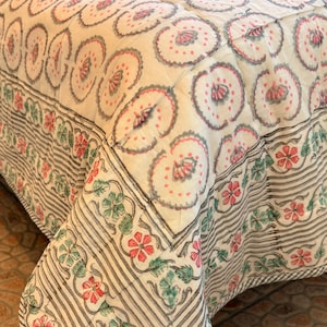 Könnte beinhalten: Eine gesteppte Tagesdecke mit weißem Untergrund und einem sich wiederholenden Muster aus kreisförmigen Blumenmotiven in gedecktem Rosa und Grau. Der Rand zeigt eine dekorative Anordnung aus rosa und grünen Blumen mit grauen Streifen.