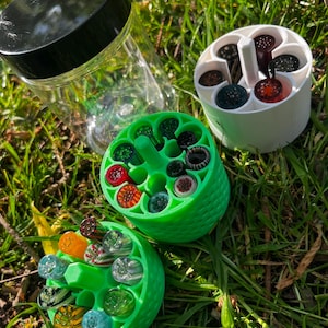 May include: A clear glass jar with a black lid is next to two green, circular containers filled with colourful glass marbles. A white container with marbles is also present. The items are arranged on green grass.