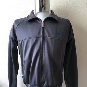 May include: Dark grey track jacket with a full zip. It has a blue Nike logo and blue detailing on the collar and sleeves. The jacket has ribbed cuffs and hem.