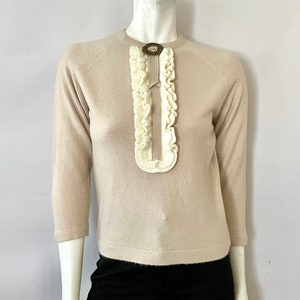 May include: A beige sweater with three-quarter sleeves and a cream-colored ruffled accent down the front. The sweater features a decorative oval-shaped brooch at the neckline. The mannequin is wearing black pants.