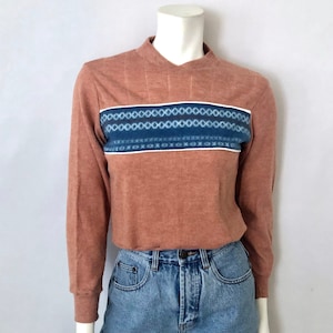 May include: A rust-colored long-sleeve shirt with a geometric design across the chest. The design features blue and white patterns. The shirt is paired with denim shorts. The outfit is displayed on a mannequin.