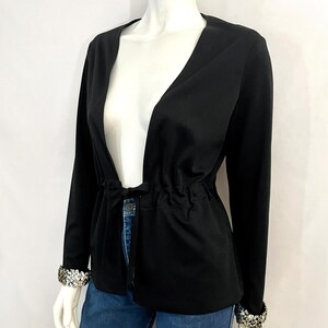 May include: A black long-sleeve jacket with a V-neck and a drawstring waist. The jacket has sequined cuffs and is displayed on a mannequin. The jacket is paired with blue jeans.