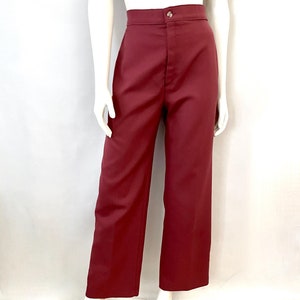 Puede incluir: Pantalones burdeos de cintura alta con cierre de botón. Los pantalones tienen un diseño de pierna ancha y se exhiben en un maniquí. La tela parece ser un material liso y sólido. Los pantalones son de color rojo oscuro.