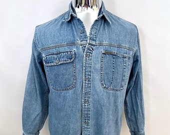 May include: Light blue denim shirt with long sleeves, two chest pockets, and a button-down front. The shirt is displayed on a mannequin against a white background. The shirt has a classic collar and a small logo on the chest pocket.