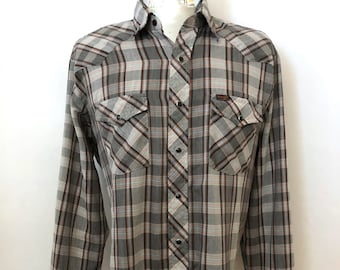 May include: A long-sleeved plaid shirt in shades of gray, brown, and red, displayed on a mannequin. The shirt features two chest pockets with button closures and a button-down front. The Wrangler brand label is visible on the pocket.
