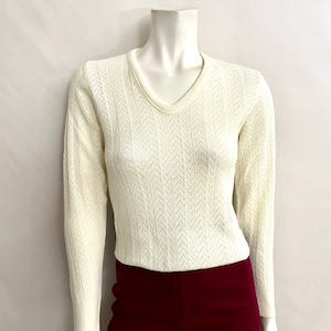 May include: Cream-colored, long-sleeved sweater with a V-neck and a delicate openwork pattern. The sweater is paired with burgundy-colored pants. The outfit is displayed on a mannequin.