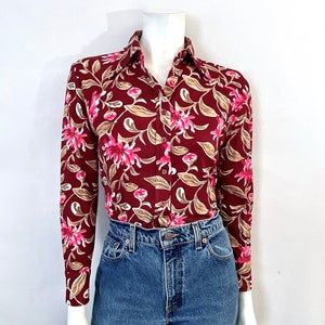 May include: A burgundy long-sleeve button-down shirt with a floral pattern of pink flowers and tan leaves. The shirt is paired with blue denim jeans. The shirt is displayed on a mannequin.