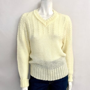 May include: A cream-colored V-neck sweater with a ribbed hem and cuffs. The sweater has a textured knit pattern on the shoulders and sleeves. The garment is displayed on a mannequin, paired with dark blue denim jeans.