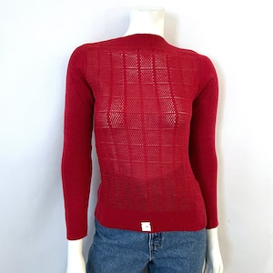 May include: A red long-sleeved sweater with a unique openwork design on the front and back. The sweater has a high neckline and is displayed on a mannequin. The bottom of the sweater has a small white tag.