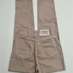May include: A pair of light brown pants featuring a high-waisted design with two back pockets and a white rectangular patch on the waistband with the word BONGO printed in black capital letters. The pants have a straight leg cut and belt loops.