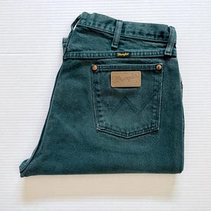 May include: Folded dark teal Wrangler jeans. The jeans feature a classic five-pocket design, belt loops, and a brown leather patch with the Wrangler logo on the back pocket. The jeans are made of a durable denim material.