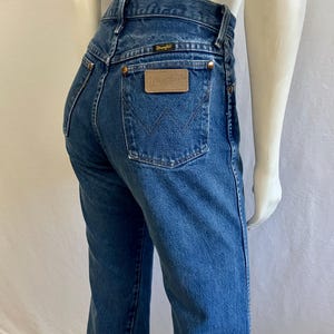 May include: Blue denim jeans with a high waist and a classic five-pocket design. The back features a leather patch with the "Wrangler" logo and a stitched "W" on the back pockets. The jeans are displayed on a mannequin.