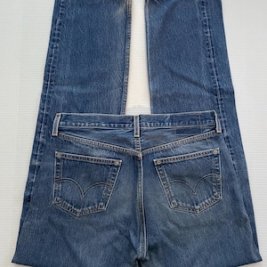 May include: A pair of blue denim jeans, laid flat. The jeans have a classic five-pocket design, with two back pockets featuring a distinctive curved stitch. The denim has a medium wash with subtle fading.