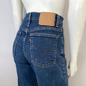 Puede incluir: Vaqueros de mezclilla azul oscuro con cintura alta y diseño clásico de cinco bolsillos. Los vaqueros presentan un parche de cuero marrón en la cinturilla trasera y la icónica etiqueta roja de Levi's en el bolsillo trasero. Los vaqueros se exhiben en un maniquí.