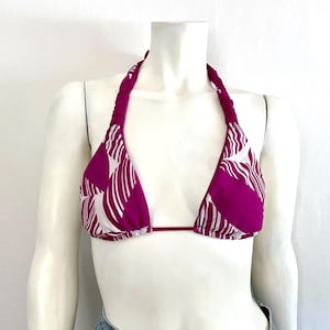 May include: A magenta halter bikini top with a white leaf pattern. The top is displayed on a mannequin. The halter straps are braided. The bikini top is a triangle shape.