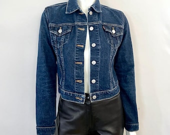 May include: A dark blue denim jacket with a classic design. The jacket features a button-up front, two chest pockets, and a collar. The jacket is displayed on a mannequin, paired with black pants.