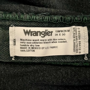 May include: Close-up of a dark gray Wrangler jeans label. The label is white with black text, including the brand name, size 36 x 30, and care instructions. The jeans are made of 100% cotton.