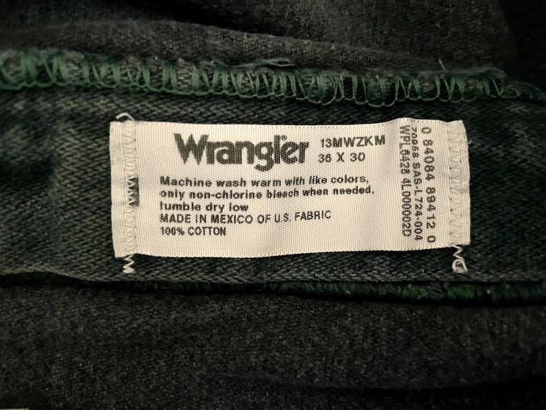 May include: Close-up of a dark gray Wrangler jeans label. The label is white with black text, including the brand name, size 36 x 30, and care instructions. The jeans are made of 100% cotton.