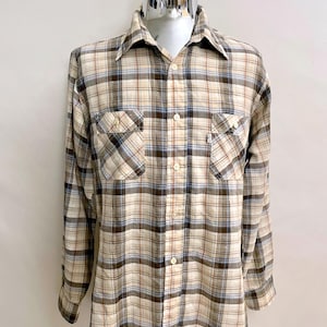 Puede incluir: Camisa de cuadros de manga larga en tonos beige, marrón y azul. La camisa tiene dos bolsillos en el pecho y una botonadura. La tela parece ligera, adecuada para ropa informal. La camisa está sobre un maniquí.