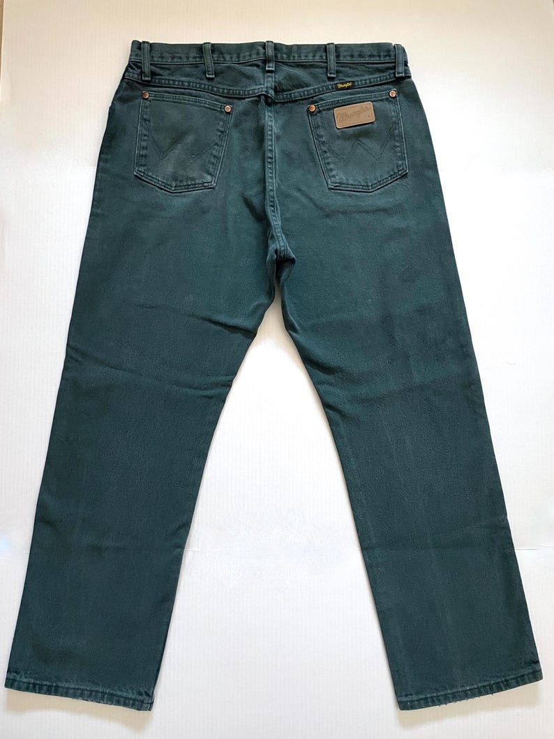 May include: Dark teal Wrangler jeans, viewed from the back, showcasing the pockets, belt loops, and a leather patch. The jeans have a classic five-pocket design and a straight leg cut. The denim appears to be a medium weight.