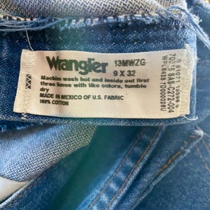 May include: Close-up of a blue denim Wrangler jeans label. The label includes the brand name, size 9 x 32, and washing instructions. The jeans are made in Mexico of U.S. fabric and are 100% cotton.
