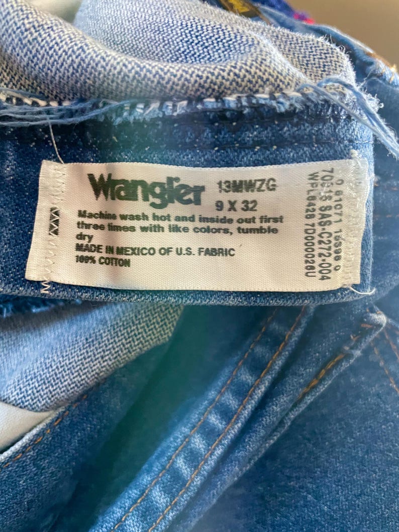 May include: Close-up of a blue denim Wrangler jeans label. The label includes the brand name, size 9 x 32, and washing instructions. The jeans are made in Mexico of U.S. fabric and are 100% cotton.