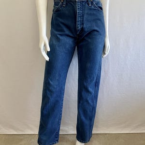 May include: Dark blue denim jeans with a button and zipper closure. The jeans have a classic five-pocket design and a straight leg fit. The jeans are displayed on a mannequin.