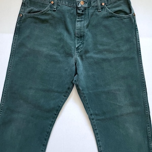 May include: Dark green denim jeans with a button and zipper closure. The jeans have classic five-pocket styling and copper-colored rivets. The fabric appears slightly faded, giving them a vintage look. The jeans are laid flat on a white surface.