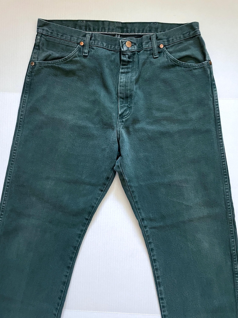 May include: Dark green denim jeans with a button and zipper closure. The jeans have classic five-pocket styling and copper-colored rivets. The fabric appears slightly faded, giving them a vintage look. The jeans are laid flat on a white surface.
