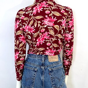 May include: A burgundy blouse with a floral pattern of pink flowers and tan leaves. The blouse is paired with blue denim jeans. The back of the blouse is visible, showing the collar and long sleeves. The jeans have a classic five-pocket design.