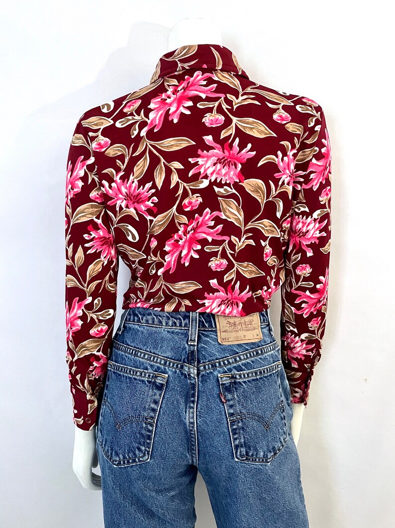 May include: A burgundy blouse with a floral pattern of pink flowers and tan leaves. The blouse is paired with blue denim jeans. The back of the blouse is visible, showing the collar and long sleeves. The jeans have a classic five-pocket design.